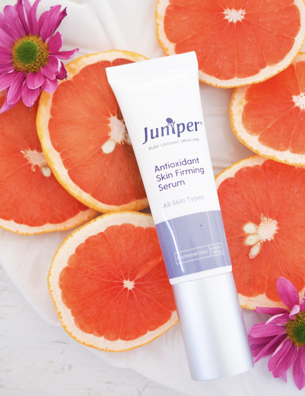 Juniper Antioxidant Skin Firming Serum tube styled among fresh grapefruit slices and pink flowers.