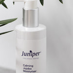 White Juniper Calming Jojoba Moisturiser pump bottle displayed on a light stone ledge beneath green olive leaves.