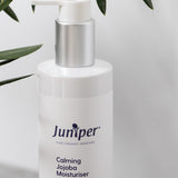 White Juniper Calming Jojoba Moisturiser pump bottle displayed on a light stone ledge beneath green olive leaves.