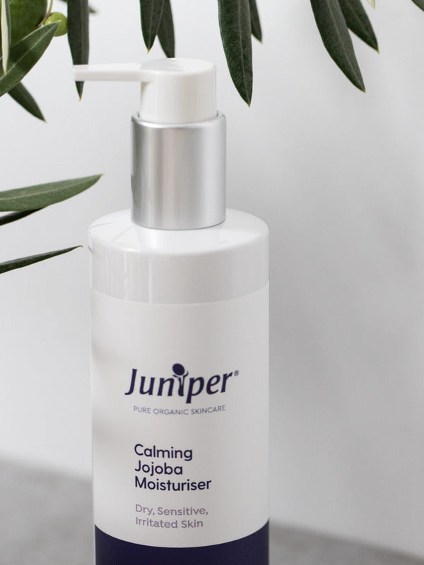 White Juniper Calming Jojoba Moisturiser pump bottle displayed on a light stone ledge beneath green olive leaves.