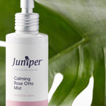 Juniper Calming Rose-Otto Mist bottle styled in front of glossy green leaves.