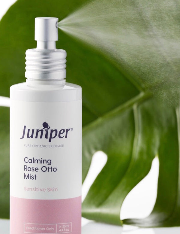 Juniper Calming Rose-Otto Mist bottle styled in front of glossy green leaves.