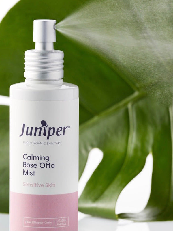 Juniper Calming Rose-Otto Mist bottle styled in front of glossy green leaves.