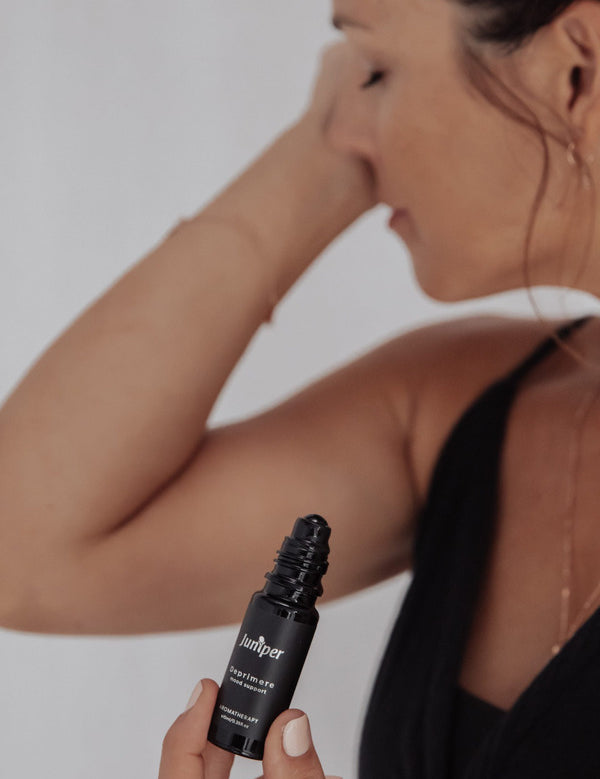 Person with eyes closed touching their forehead with one hand while holding an uncapped black Juniper Deprimere roll-on bottle in the other, wearing a black sleeveless top.