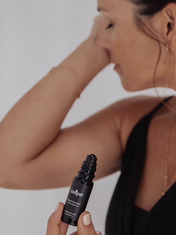 Person with eyes closed touching their forehead with one hand while holding an uncapped black Juniper Deprimere roll-on bottle in the other, wearing a black sleeveless top.