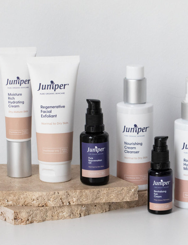 Seven Juniper mature skincare products, including tubes, pump bottles and dark glass treatment bottles, arranged on a natural stone platform.