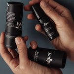 Hands holding a capped black Juniper Studium roll-on bottle beside its open cylindrical black cardboard packaging, with white leaf illustrations and the detached outer cover resting across the palm.