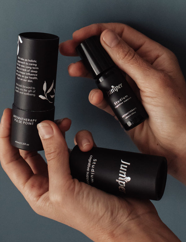Hands holding a capped black Juniper Studium roll-on bottle beside its open cylindrical black cardboard packaging, with white leaf illustrations and the detached outer cover resting across the palm.