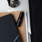 Juniper Studium 10ml aromatherapy roller, included in the Cognitive Support + Wellbeing Collection, resting on notebooks beside a pen and wristwatch.