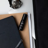 Juniper Studium 10ml aromatherapy roller, included in the Cognitive Support + Wellbeing Collection, resting on notebooks beside a pen and wristwatch.