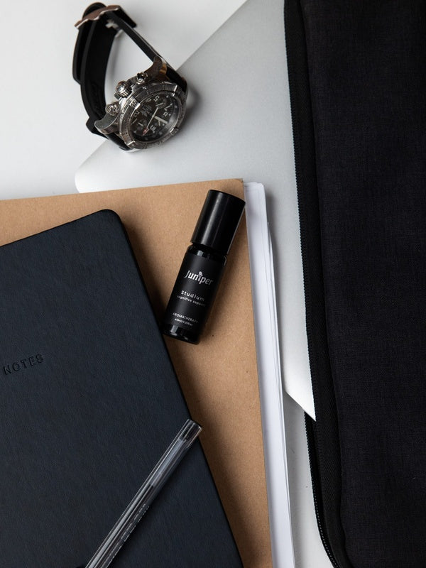 Juniper Studium 10ml aromatherapy roller, included in the Cognitive Support + Wellbeing Collection, resting on notebooks beside a pen and wristwatch.