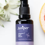 Juniper Vitamin C Night Serum bottle styled with white flowers and a fresh grapefruit slice.