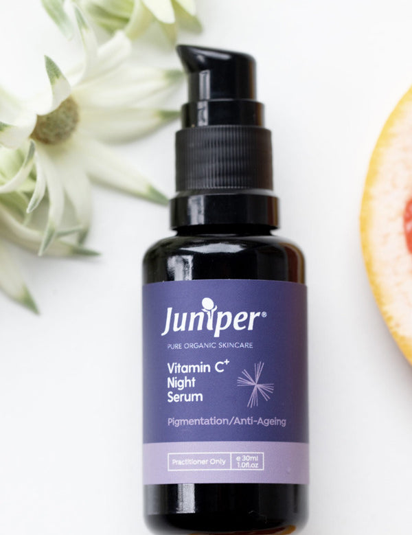 Juniper Vitamin C Night Serum bottle styled with white flowers and a fresh grapefruit slice.