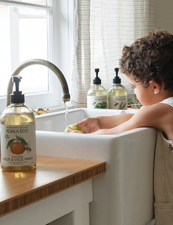 Child washing a green apple under running water in a white kitchen sink. A Koala Eco Fruit & Vege Wash bottle stands on the wooden counter in front, with two more pump bottles beside the window.