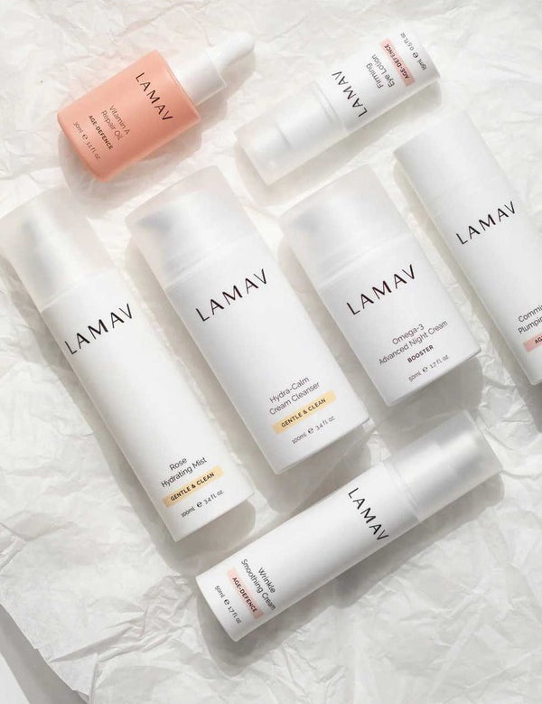 Seven white and blush LAMAV Age-Defence skincare products for dry and sensitive skin arranged diagonally on crumpled white paper.