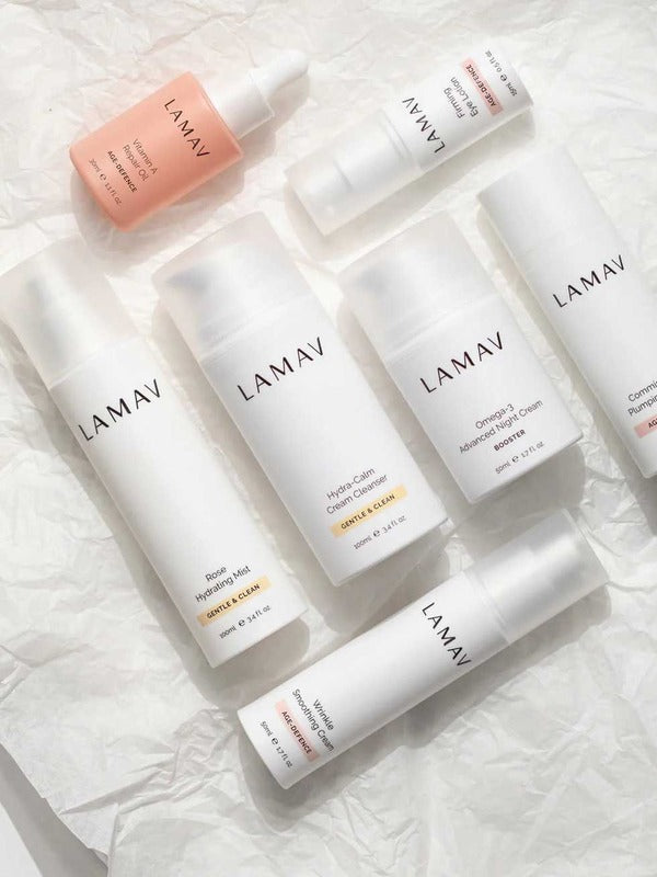 Seven white and blush LAMAV Age-Defence skincare products for dry and sensitive skin arranged diagonally on crumpled white paper.