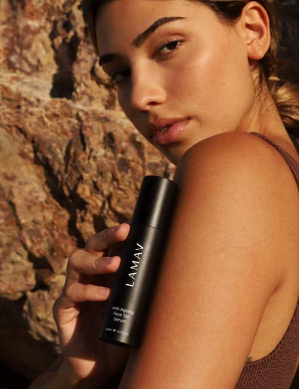 A woman with radiant skin stands outdoors against rocks, wearing a sleeveless brown top and holding a black bottle of LAMAV Anti-Ageing Face Tan Serum 50ml near her face.