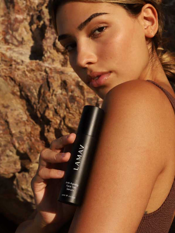 A woman with radiant skin stands outdoors against rocks, wearing a sleeveless brown top and holding a black bottle of LAMAV Anti-Ageing Face Tan Serum 50ml near her face.
