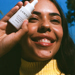 Smiling woman outdoors holding a bottle of LAMAV Bakuchiol 4% Natural Retinol Alternative Booster beside her face in bright natural light.