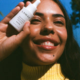 Smiling woman outdoors holding a bottle of LAMAV Bakuchiol 4% Natural Retinol Alternative Booster beside her face in bright natural light.