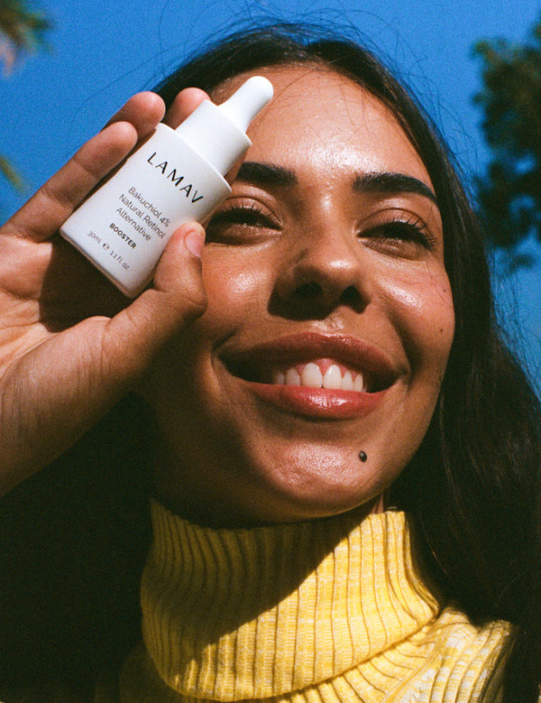 Smiling woman outdoors holding a bottle of LAMAV Bakuchiol 4% Natural Retinol Alternative Booster beside her face in bright natural light.