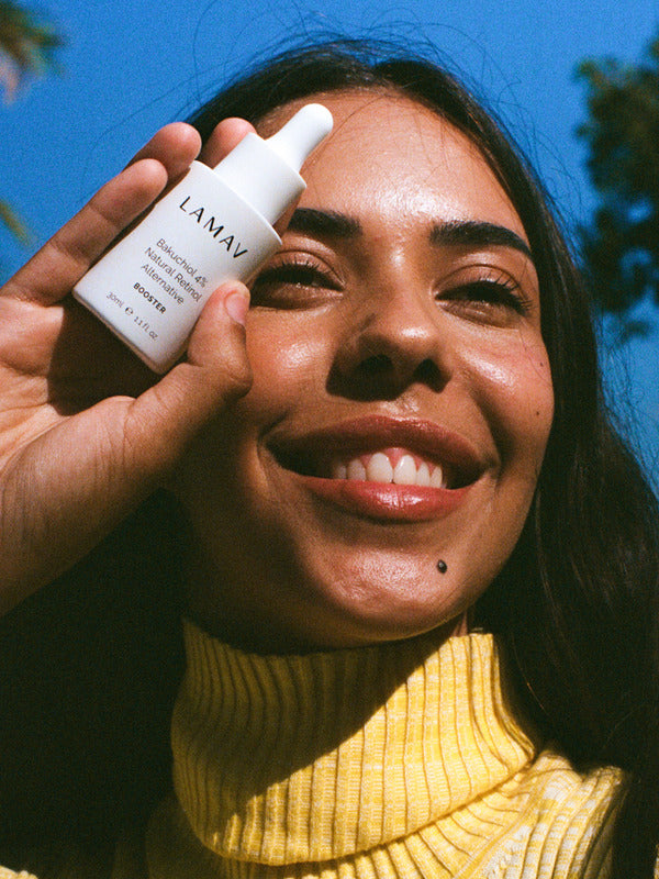Smiling woman outdoors holding a bottle of LAMAV Bakuchiol 4% Natural Retinol Alternative Booster beside her face in bright natural light.