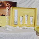 An open yellow LAMAV Brightening box displays one tall white bottle and three small white tubes in fitted recesses on draped white fabric. Its lid, showing a woman's face, stands behind it.
