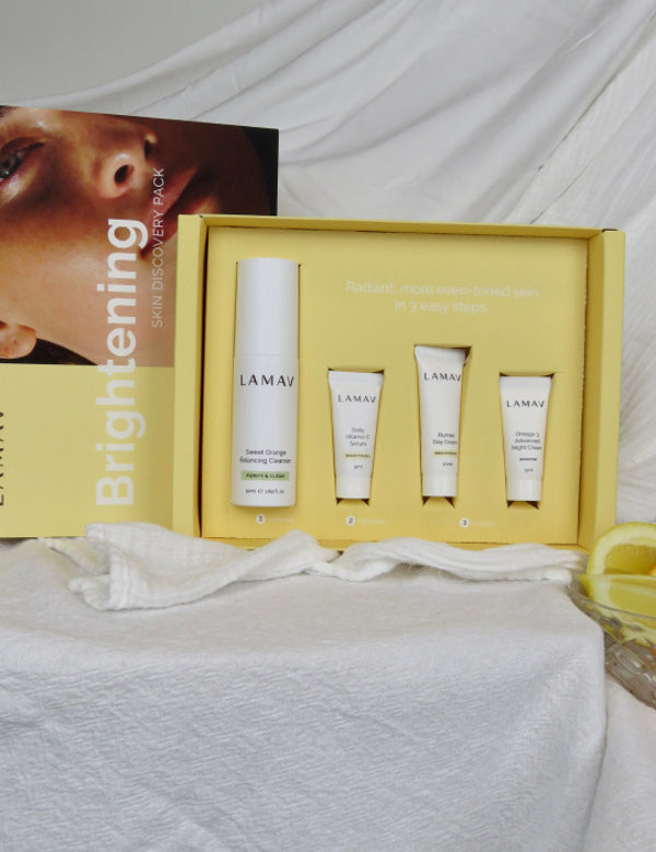 An open yellow LAMAV Brightening box displays one tall white bottle and three small white tubes in fitted recesses on draped white fabric. Its lid, showing a woman's face, stands behind it.