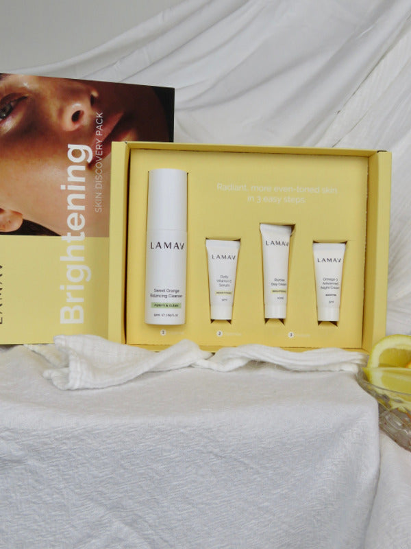 An open yellow LAMAV Brightening box displays one tall white bottle and three small white tubes in fitted recesses on draped white fabric. Its lid, showing a woman's face, stands behind it.