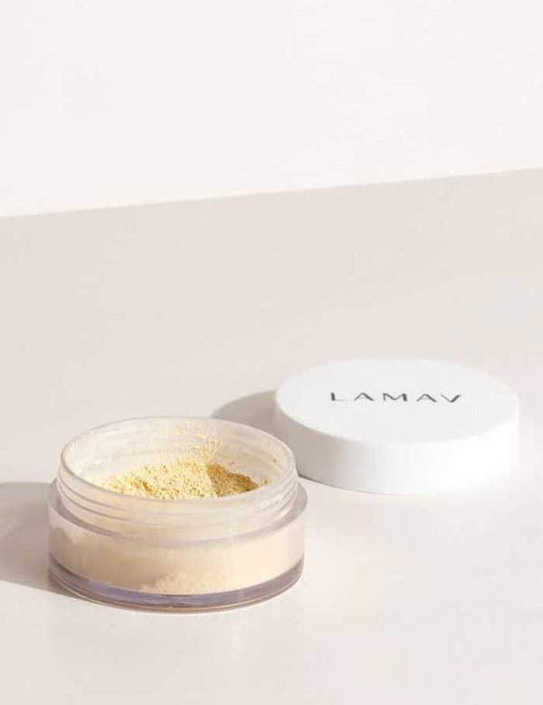 An open LAMAV Mattifying Powder 3g jar with a white lid rests on a light surface. The clear container reveals some of the yellow-tinted, talc-free powder inside.