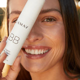 Smiling woman holding a white LAMAV Certified Organic BB Cream tube beside her face in warm natural light.