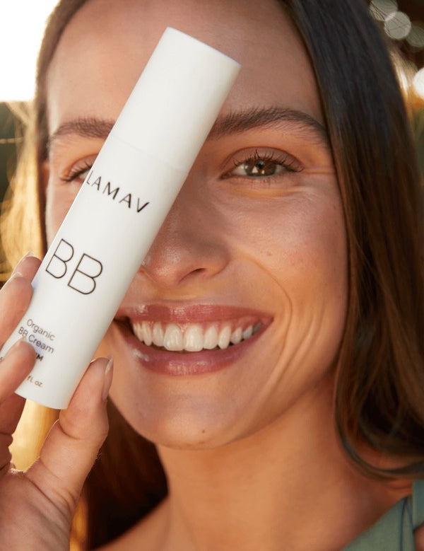 Smiling woman holding a white LAMAV Certified Organic BB Cream tube beside her face in warm natural light.