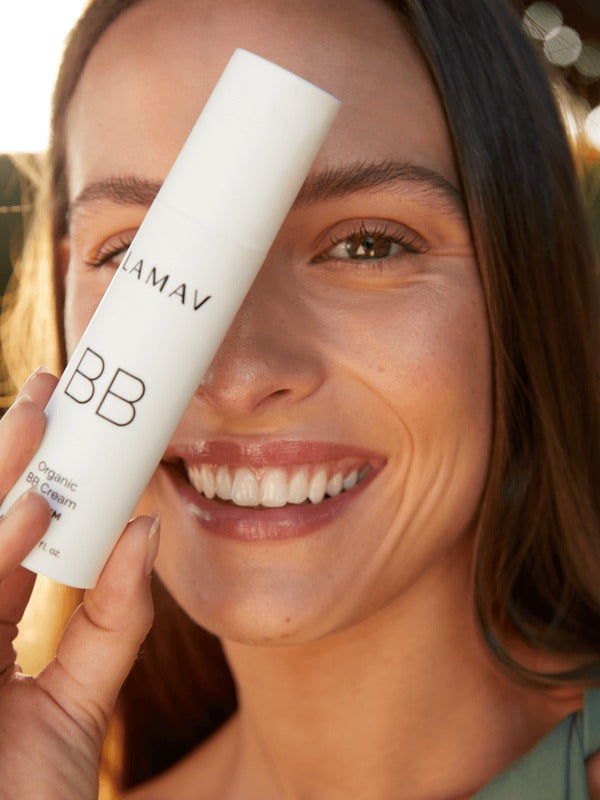 Smiling woman holding a white LAMAV Certified Organic BB Cream tube beside her face in warm natural light.