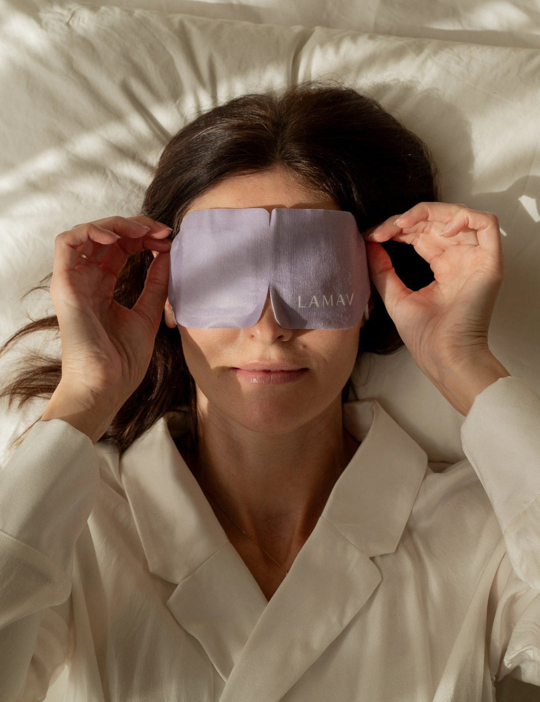 A woman lies on cream bedding and holds the sides of the purple LAMAV Lavender Self Warming Eye Mask over her closed eyes, demonstrating how the mask is worn.