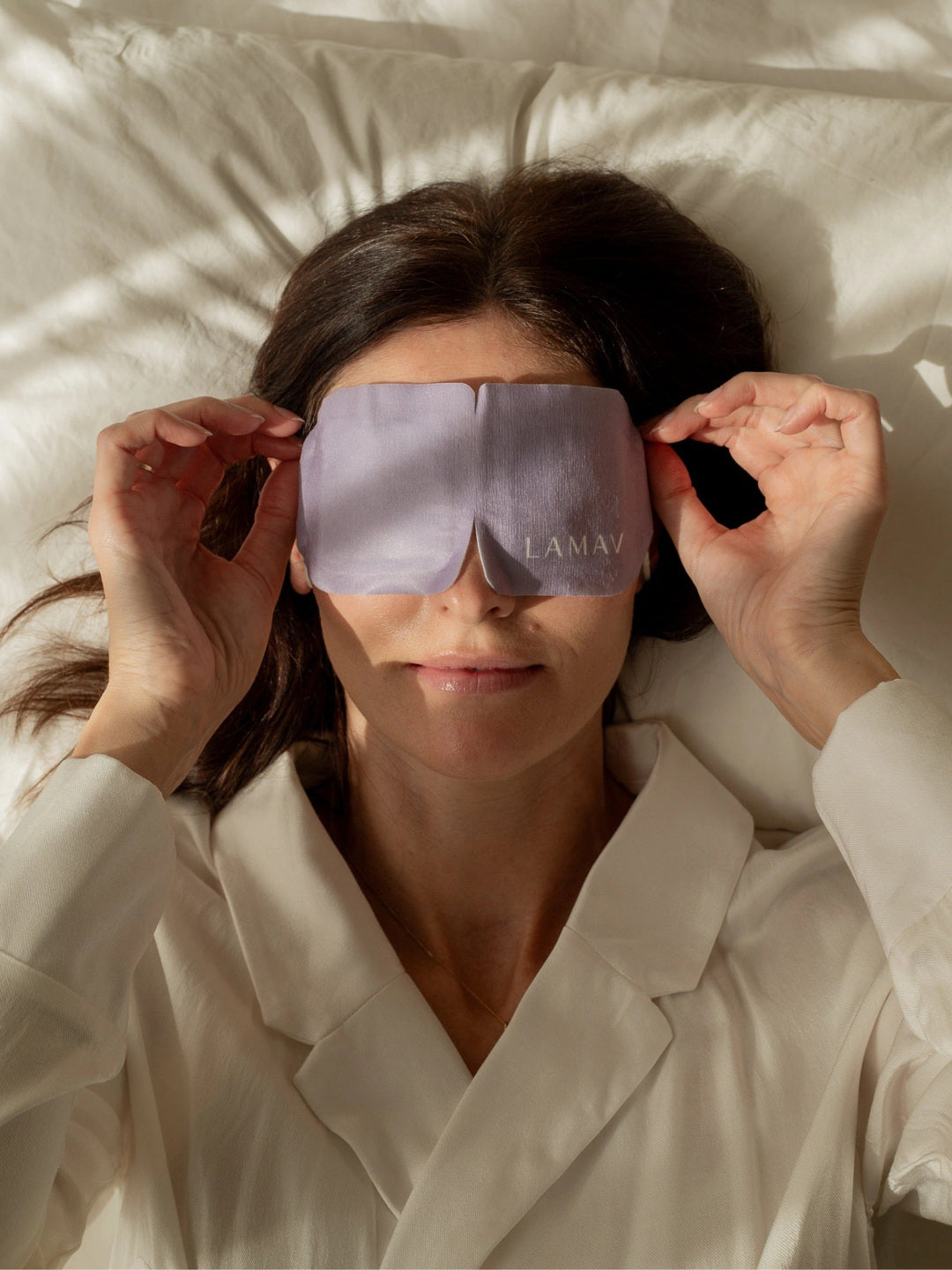 A woman lies on cream bedding and holds the sides of the purple LAMAV Lavender Self Warming Eye Mask over her closed eyes, demonstrating how the mask is worn.