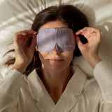 A woman lies on cream bedding and holds the sides of the purple LAMAV Lavender Self Warming Eye Mask over her closed eyes, demonstrating how the mask is worn.