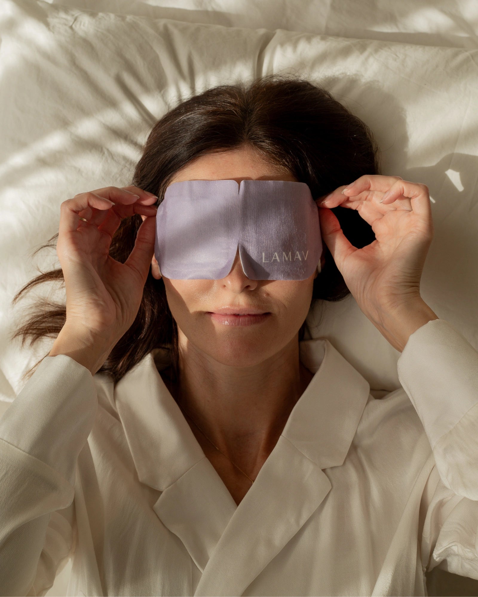 A woman lies on cream bedding and holds the sides of the purple LAMAV Lavender Self Warming Eye Mask over her closed eyes, demonstrating how the mask is worn.
