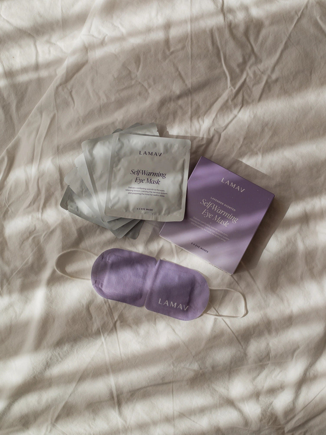 LAMAV Lavender Self Warming Eye Mask displayed with its purple five-mask box, silver individual sachets and white ear loops on softly folded cream bedding.