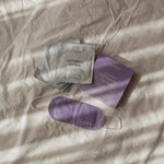 LAMAV Lavender Self Warming Eye Mask displayed with its purple five-mask box, silver individual sachets and white ear loops on softly folded cream bedding.
