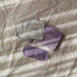 LAMAV Lavender Self Warming Eye Mask displayed with its purple five-mask box, silver individual sachets and white ear loops on softly folded cream bedding.