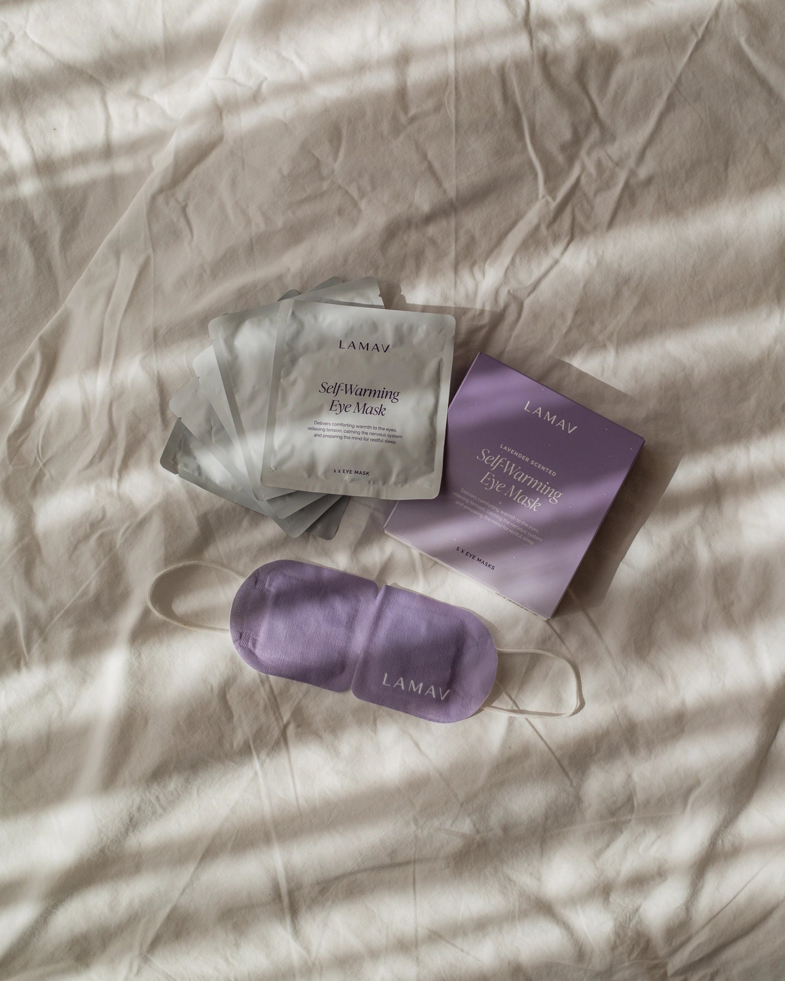 LAMAV Lavender Self Warming Eye Mask displayed with its purple five-mask box, silver individual sachets and white ear loops on softly folded cream bedding.