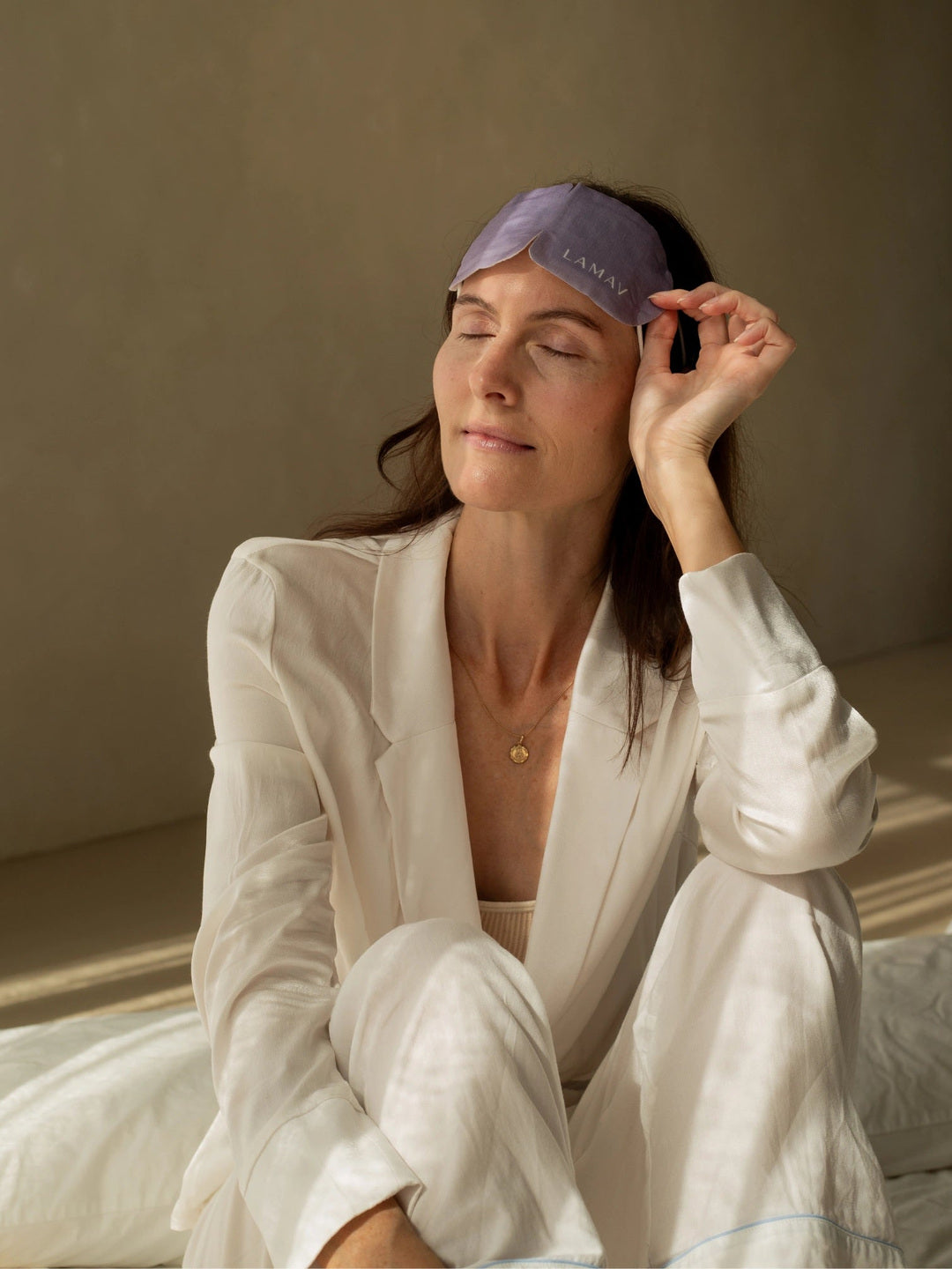 A woman seated on a bed lifts the purple LAMAV Lavender Self Warming Eye Mask onto her forehead, holding one side to show how it is removed after use.