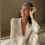 A woman seated on a bed lifts the purple LAMAV Lavender Self Warming Eye Mask onto her forehead, holding one side to show how it is removed after use.