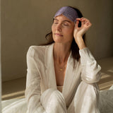 A woman seated on a bed lifts the purple LAMAV Lavender Self Warming Eye Mask onto her forehead, holding one side to show how it is removed after use.