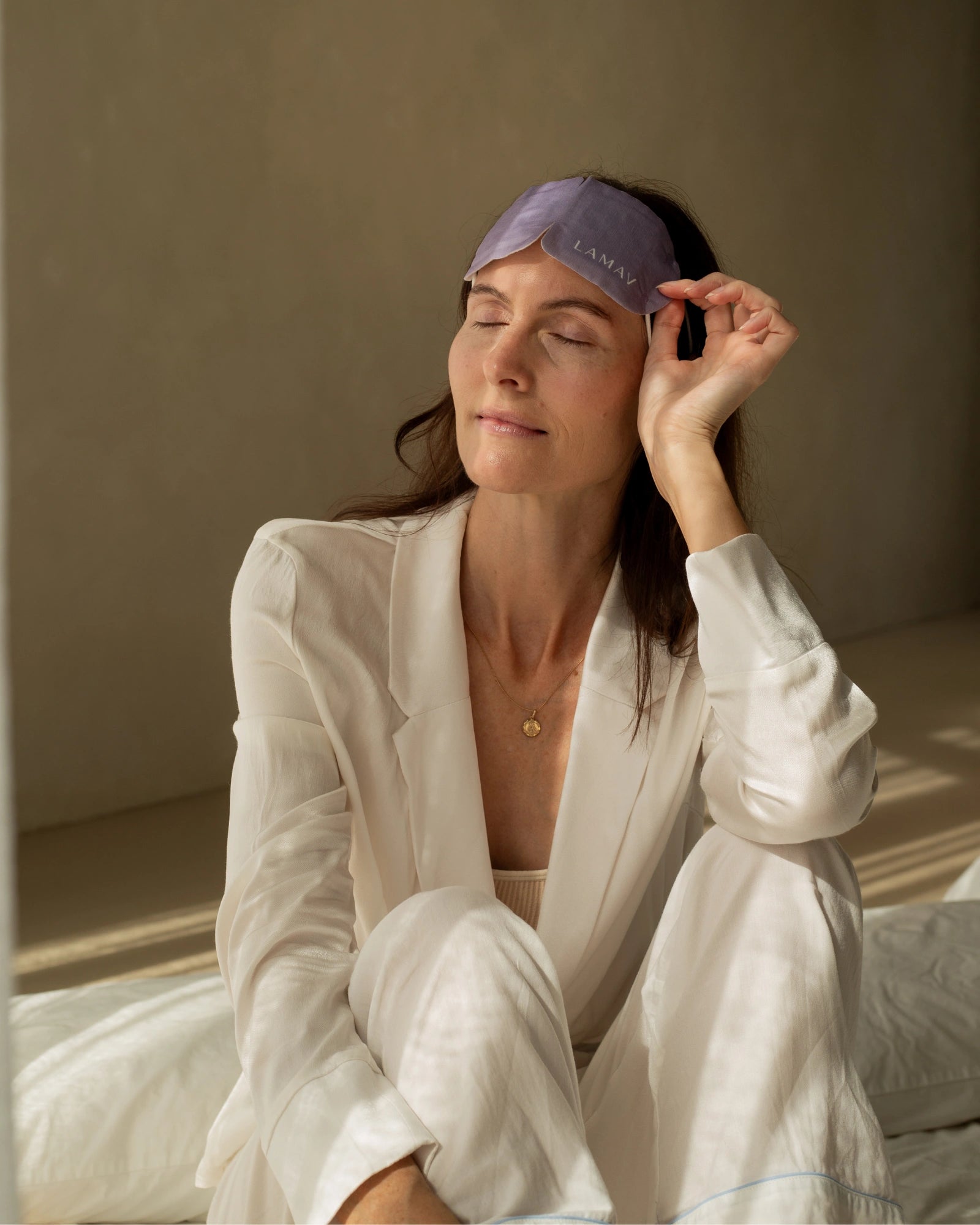 A woman seated on a bed lifts the purple LAMAV Lavender Self Warming Eye Mask onto her forehead, holding one side to show how it is removed after use.
