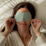 A woman reclines on cream bedding while adjusting the mint-green LAMAV Unscented Self Warming Eye Mask over her eyes, with both hands holding the lower edges.