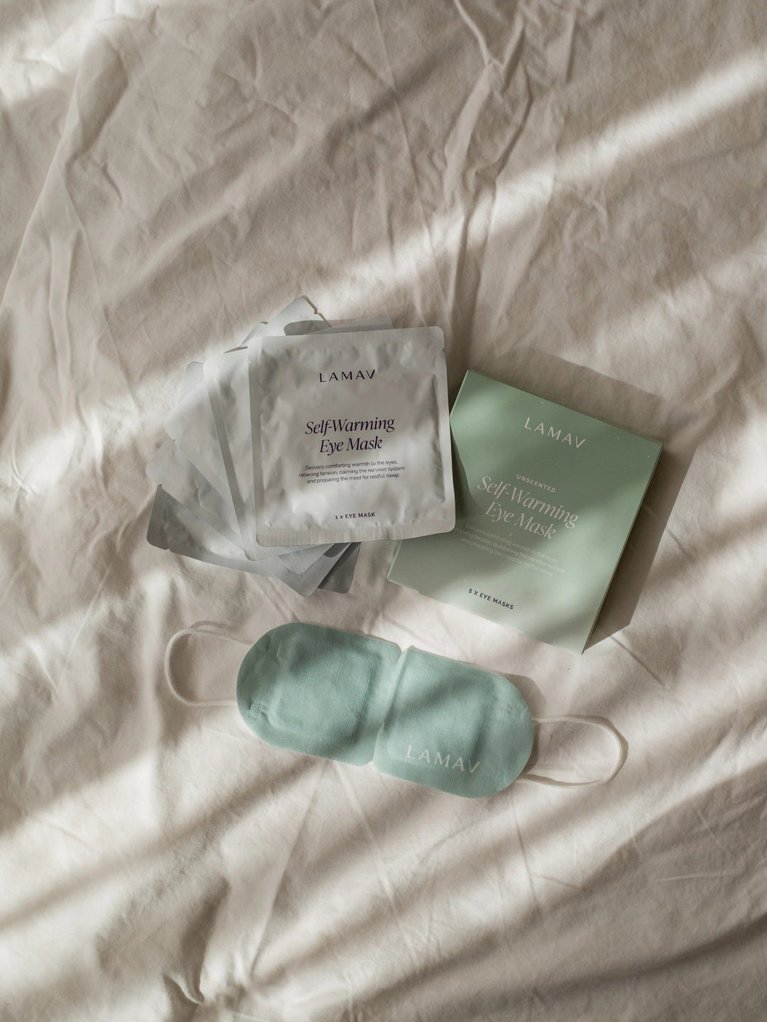LAMAV Unscented Self Warming Eye Mask with its green five-mask box and individually sealed silver sachets arranged on cream bedding; the green mask and white ear loops are visible.