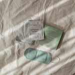 LAMAV Unscented Self Warming Eye Mask with its green five-mask box and individually sealed silver sachets arranged on cream bedding; the green mask and white ear loops are visible.