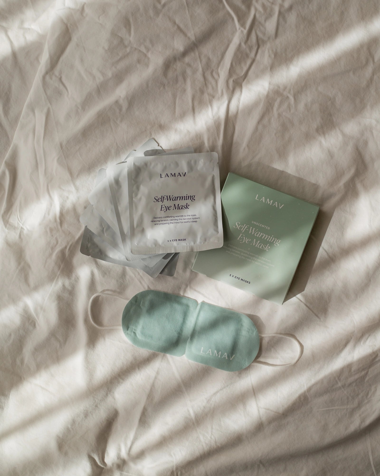 LAMAV Unscented Self Warming Eye Mask with its green five-mask box and individually sealed silver sachets arranged on cream bedding; the green mask and white ear loops are visible.