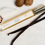 Lavanila Pure Vanilla rollerball perfume with gold cap arranged on pale wood beside vanilla pods, an orchid and tonka beans.